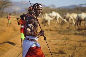 Ein Samburu-Krieger in traditioneller Kleidung trägt einen Speer, im Hintergrund sind weitere Menschen sowie Kamele zu erkennen.