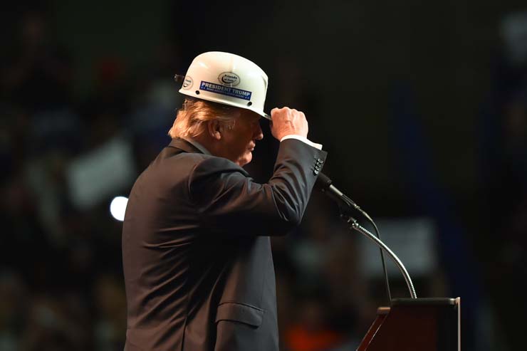 Donald Trump, an einem Mikrofon stehend, schiebt sich den Schirm eines Arbeitshelms tiefer in die Stirn.