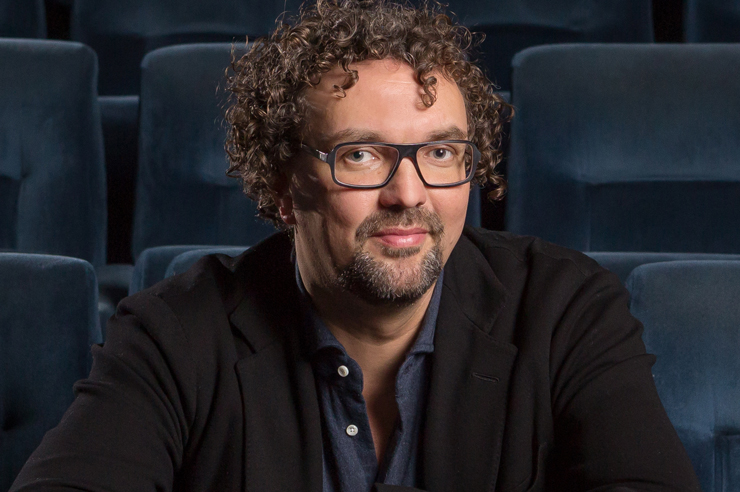 Filmemacher Trimpop mit Locken und großer Brille sitzt in einem leeren Kinosaal und blickt in die Kamera.