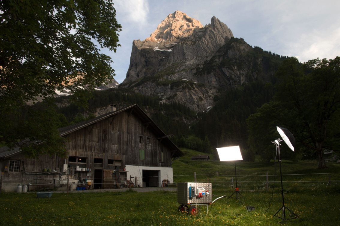 Ein alpiner Stall liegt bereits im Schatten, während eine jäh aufragende Bergspitze dahinter noch in der Sonne liegt; vor dem Gebäude wird ein Salzwasserbatterie-Anhänger mit zwei Fotolampen angeleuchtet.