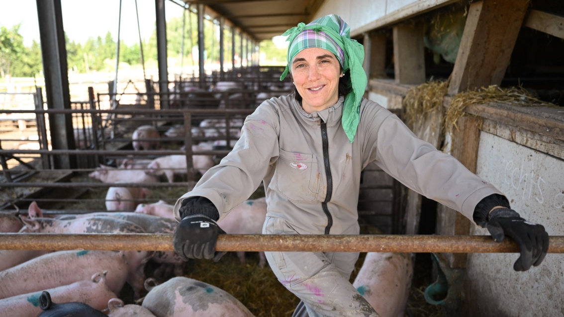Eine lächelnde Bäuerin im grauen Overall und grünem Kopftuch im Freigehege bei den Schweinen.