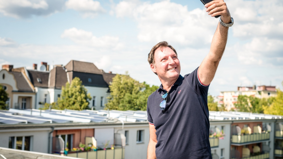 Auf einem Flachdach steht ein Mann mittleren Alters. Er hält mit der linken Hand  sein Smartphone hoch, um ein Selfie zu schießen.
