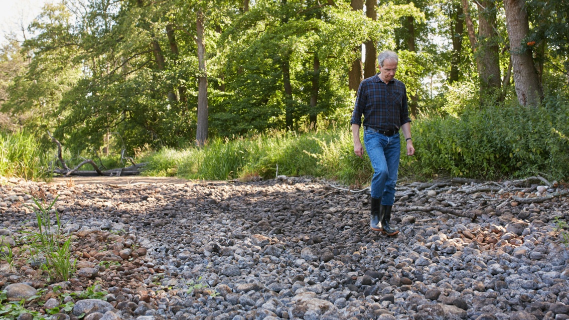 Ein Mann in Karohemd und Jeans läuft in einem ausgetrockneten Fließgraben.über Kieselsteine.