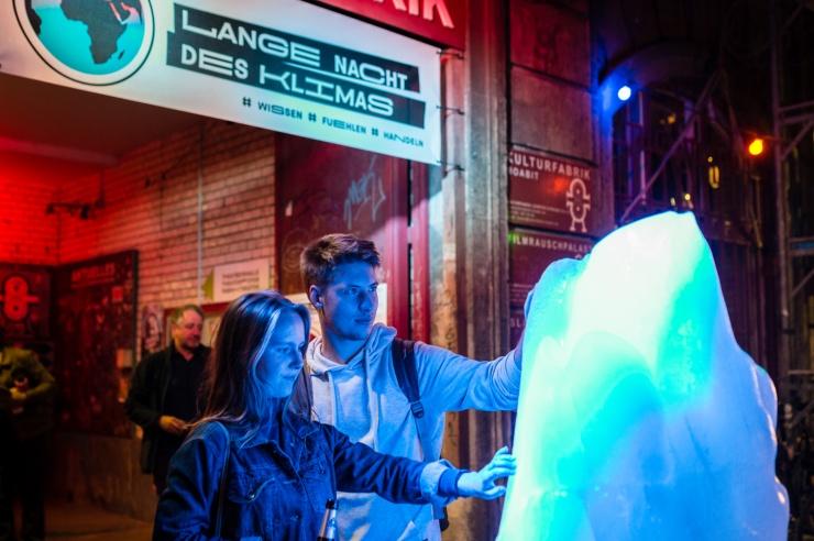 Zwei junge Menschen berühren einen wie ein Eisberg geformten Eisklotz vor dem Eingangsportal zur Langen nacht des Klimas