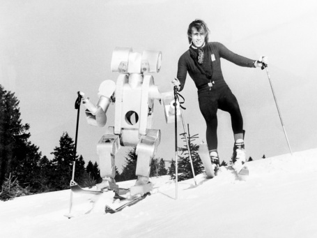 Ein junger Mann und ein gleich großer Roboter fahren auf Skiern einen Hang hinunter.