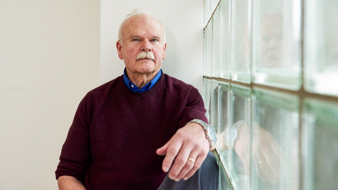 Portrait eines älteren Mannes mit Schnauzbart, der ernst in die Kamera schaut; rechts von ihm eine Wand aus Glasbausteinen.