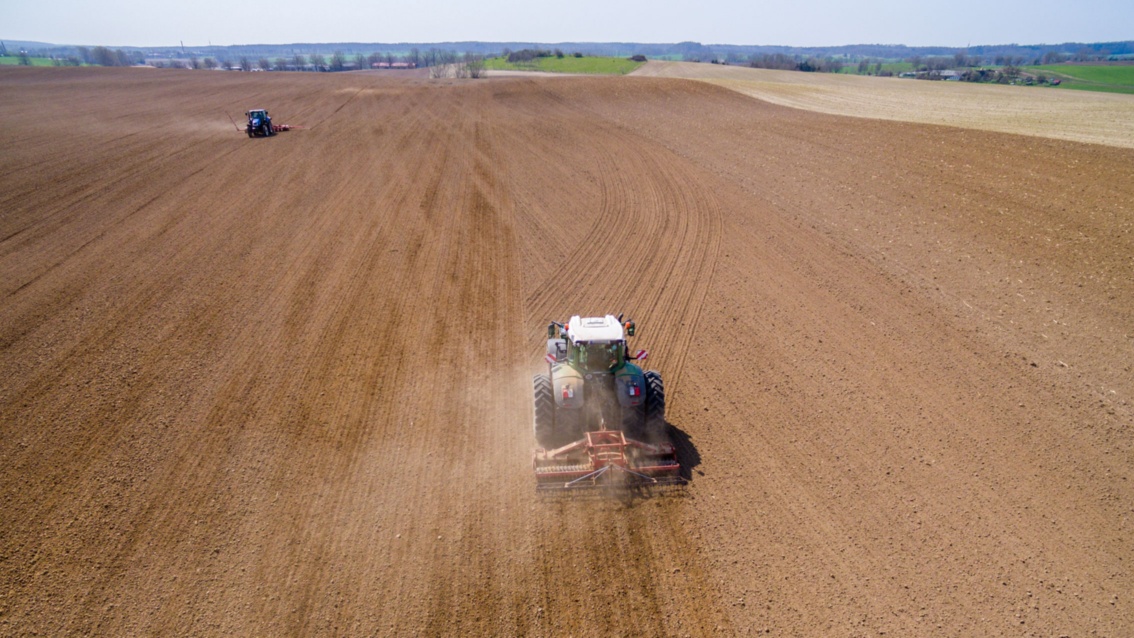 Eine weite Agrarlandschaft, auf der 2 Traktoren den trockenen Boden pflügen.