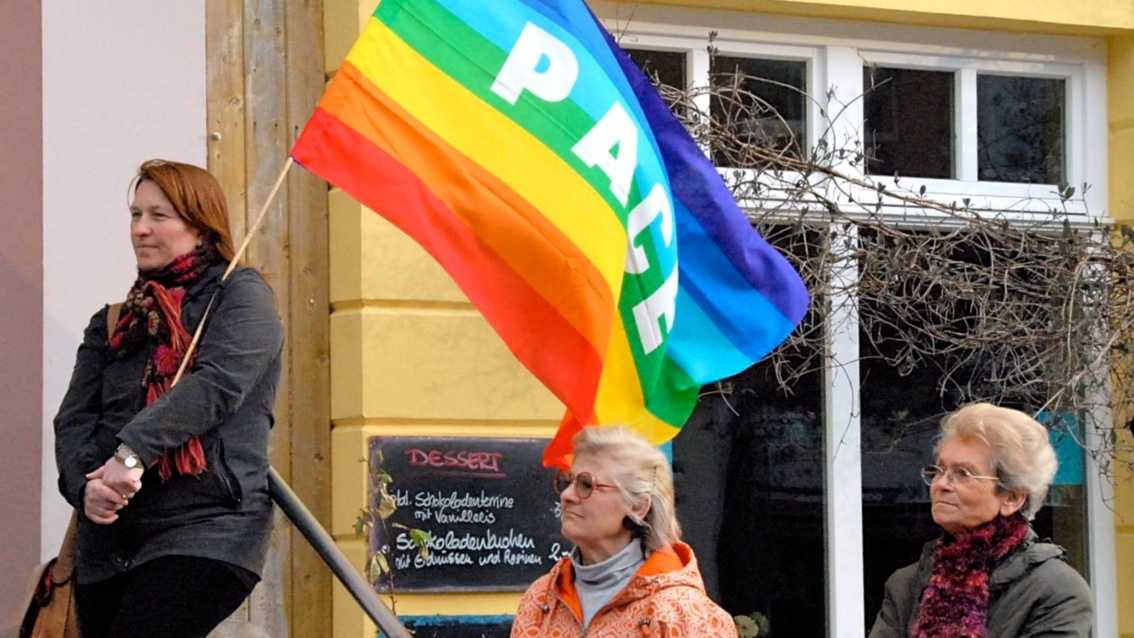 Eine Frau steht an eine Hauswand gelehnt, stolz. Sie trägt eine regenbogenfarbene Fahne mit dem Aufdruck «Peace». 