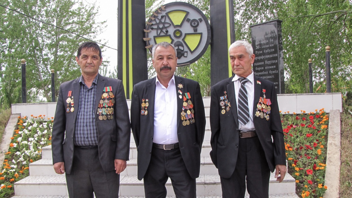 Vor einem Denkmal mit kyrillischer Inschrift stehen drei alte Männer in Anzügen, die mit zahlreichen Orden behangen sind.