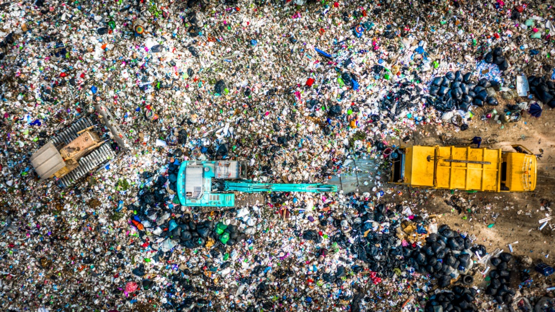 Auf einer großen Mülldeponie, die von oben fotografiert wird, steht ein blauer Bagger und ein gelbes Müllfahrzeug.