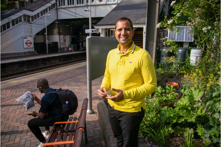 A friendly smiling man in a yellow jumper stands on a London Underground platform.
