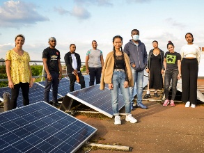 A group of young people of different skin colors form a group, with several solar panels between them.