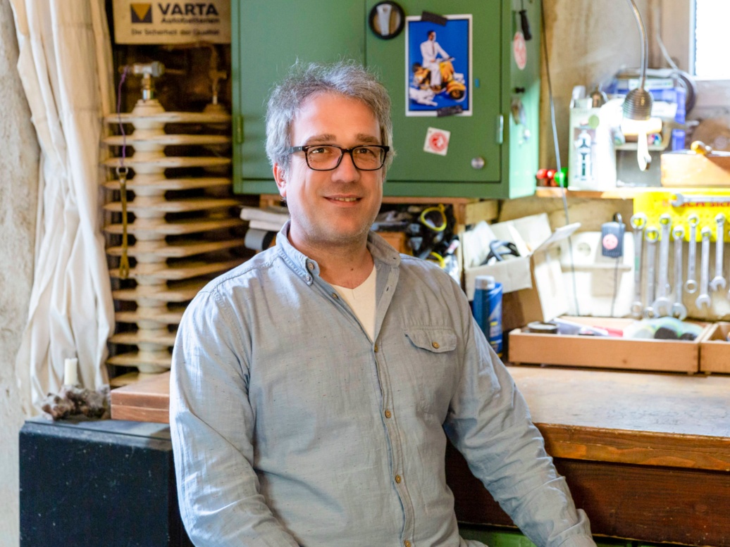 In einer Kellerwerkstatt sitzt ein Mann mit grauen Haaren in Jeans an einem aufgeräumten Werktisch und schaut in die Kamera.