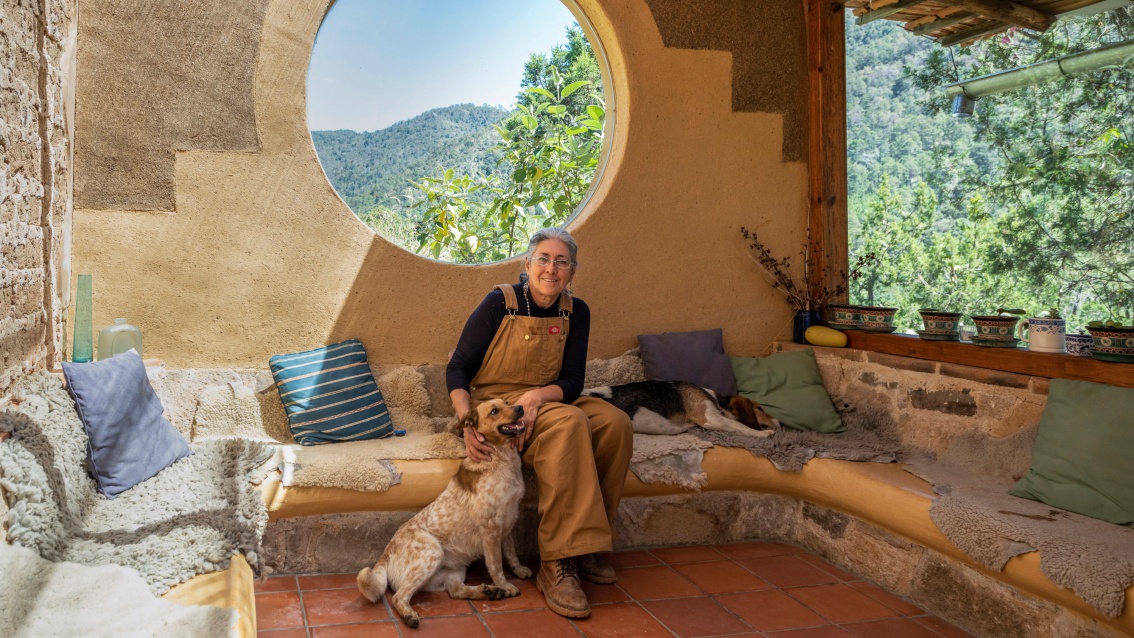 Die Architektin sitzt mit ihren beiden Hunden in einer großzügigen Veranda. Durch große runde und eckige Wanddurchbrüche schaut man in die wilde grüne Natur.