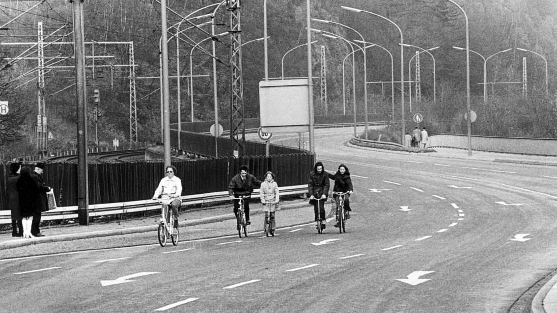 Schwarzweißfoto: Auf einer Autobahnbrücke befährt ein junger Mann mitz seinem Klapprad eine Abbiegespur; hinter ihm zwei junge Väter auf Fahrrädern, die beide ihre Kinder auf Rollern anschieben. Im Vordergrund links ein älteres Pärchen, das die Szenerie bobachtet.