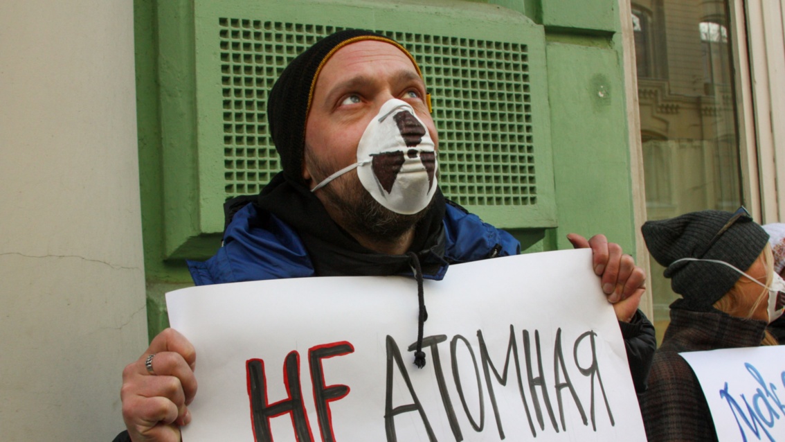 Slivyak mit weiteren Aktivisten auf der Straße: Er trägt Mütze und Atemschutz mit aufgemaltem Atomwarnzeichen, vor der Brust hält er ein Plakat. 