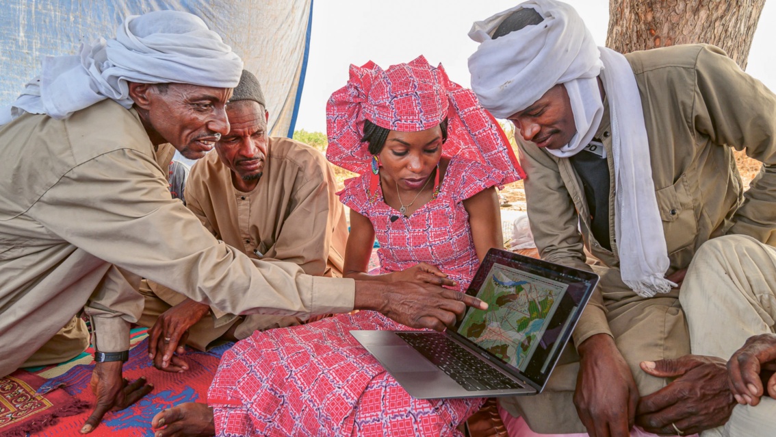Eine Frau sitzt in farbenfroher Tracht zwischen drei Männern, Beduinen, gemeinsam betrachten sie eine Karte auf ihrem Laptop. 