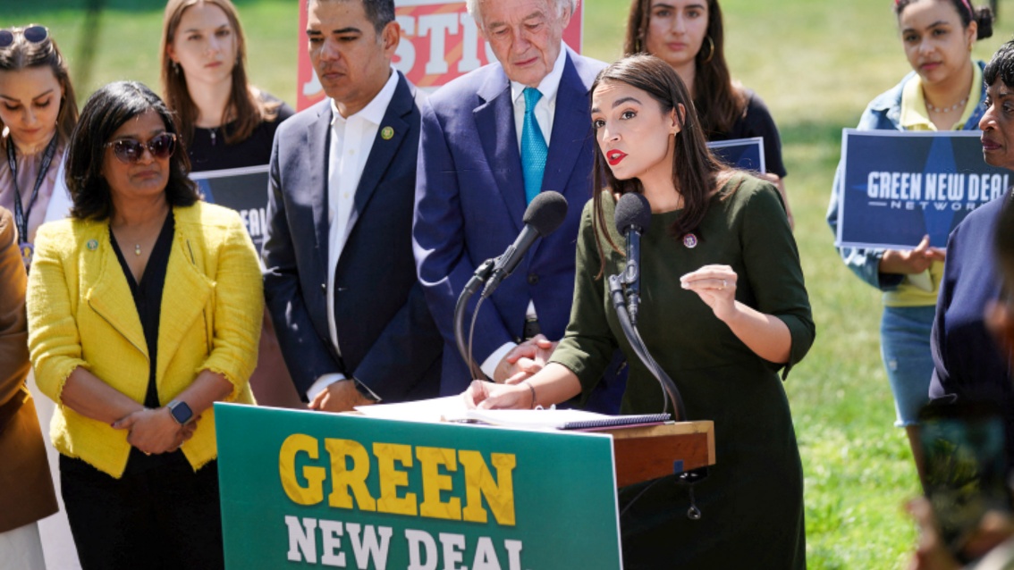 Umringt von Zuhörern, spricht eine junge Frau an einem Rednerpult, an dem ein Schild mit der Aufschrift Green New Deal hängt, im Freien eine Rede.