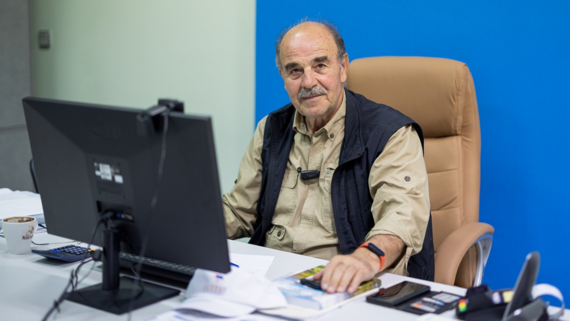 An older man sits at a desk with a PC and looks benevolently towards the camera.
