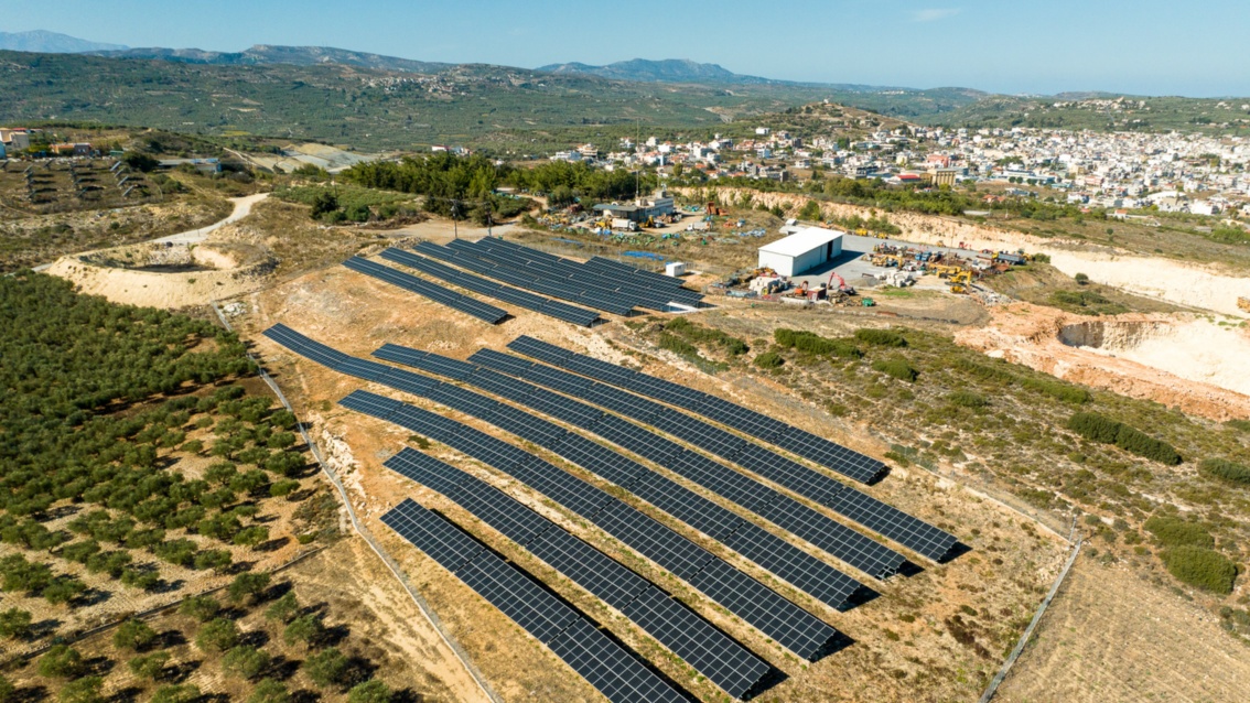 Panorama: A ground-mounted PV system is located in a wide plain between olive tree plantations and houses.