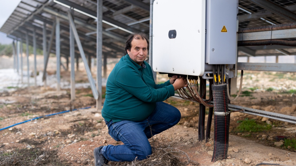 A middle-aged man in jeans and a T-shirt kneels in front of an inverter while he adjusts some cables.