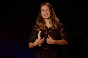 Eine junge Frau mit langen dunkelblonden Haare steht gestikulierend auf der Bühne, während sie zum Publikum spricht. 