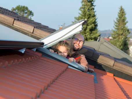 Ein Mann und ein Kind schauen fröhlich aus einem Dachfenster, vor ihnen eine PV-Dachanlage