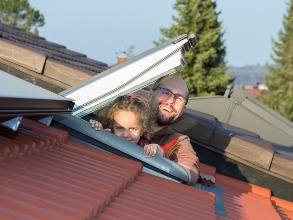 Ein Mann und ein Kind schauen fröhlich aus einem Dachfenster, vor ihnen eine PV-Dachanlage