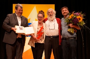 Auf einer Bühne stehen ein Mann mit einem Buch, eine Frau mit Trophäe in der Hand, ein älterer bärtiger Herr und ein Mann mit einem großen Blumenstrauß im Arm