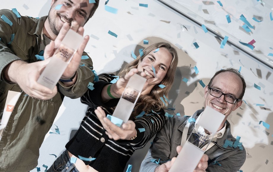 Drei Menschen stehen im Konfettiregen und halten gläserne Trophäen hoch