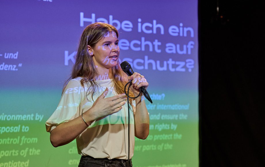 Junge Frau mit Mikrofon steht vor einer Leinwand, von der Projektion teils eingefärbt.