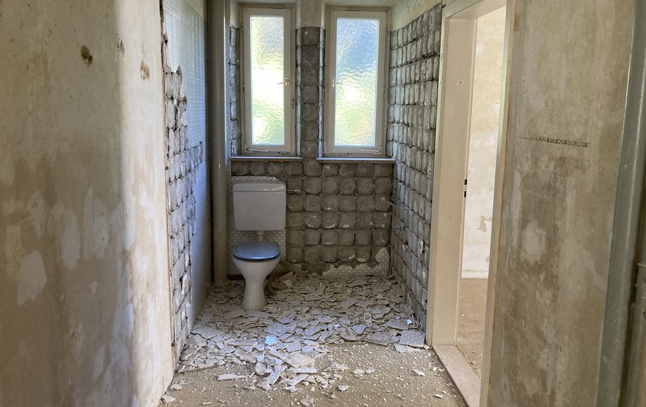 Ein altes Klo steht in einem Raum mit teils nackten Betonwänden, auf dem Boden liegen die Scherben der alten Wandfliesen