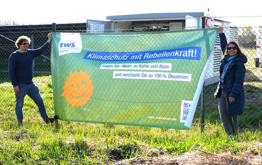 Zwei Menschen halten ein großes Banner an einen Maschendrahtzaun