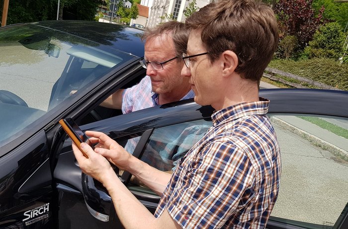 Zwei Menschen am Auto, beide gucken ins Handy