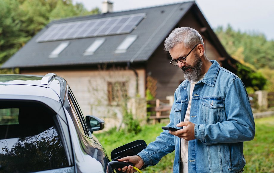 Mann lädt sein E-Auto und blickt auf sein Smartphone, im Hintergrund ein Haus mit Solardach