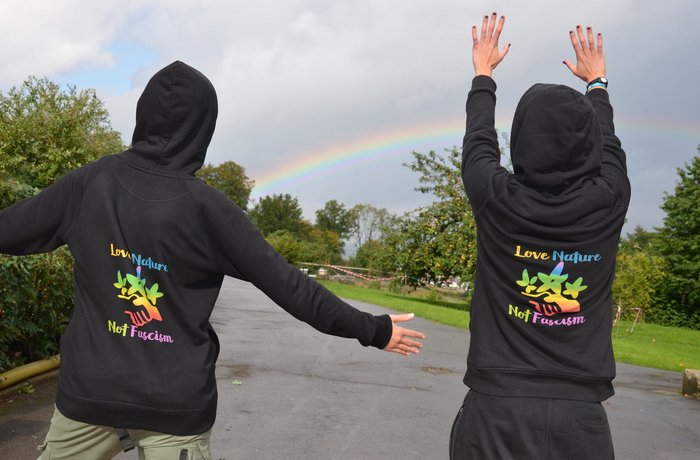 Zwei Jugendliche von hinten vor Regenbogen, auf ihren Hoodies ist der Schriftzug „Love nature, hate fascism“ zu sehen 