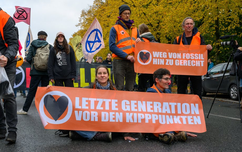 Demonstrationszug, im Vordergrund sitzen ein Mann und eine Frau auf der Straße und halten ein Plakat mit der Aufschrift "Letzte Generation vor den Kipppunkten"