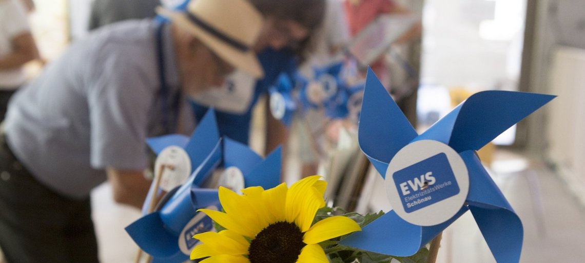 Mehrere Windräder aus Papier mit EWS-Logo und eine Sonnenblume stehen auf einem Tisch. Dahinter eine Gruppe von Menschen beim Schönauer Stromseminar 2024.