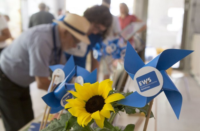 Mehrere Windräder aus Papier mit EWS-Logo und eine Sonnenblume stehen auf einem Tisch. Dahinter eine Gruppe von Menschen beim Schönauer Stromseminar 2024.