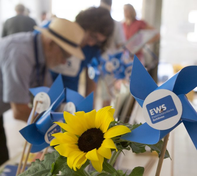 Mehrere Windräder aus Papier mit EWS-Logo und eine Sonnenblume stehen auf einem Tisch. Dahinter eine Gruppe von Menschen beim Schönauer Stromseminar 2024.