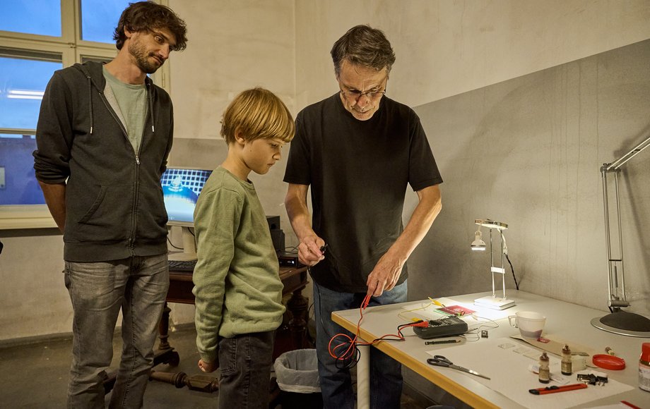 Ein Mann beugt sich über einen Tisch mit diversen Gerätschaften und hält ein Potentiometer in der Hand, daneben stehen ein Junge und sein Vater und sehen zu.
