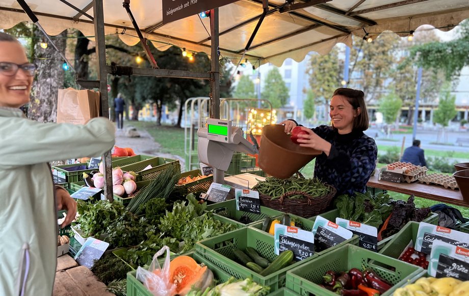 Eine junge Frau am Marktstand reicht eine Tüte Gemüse an eine Kundin