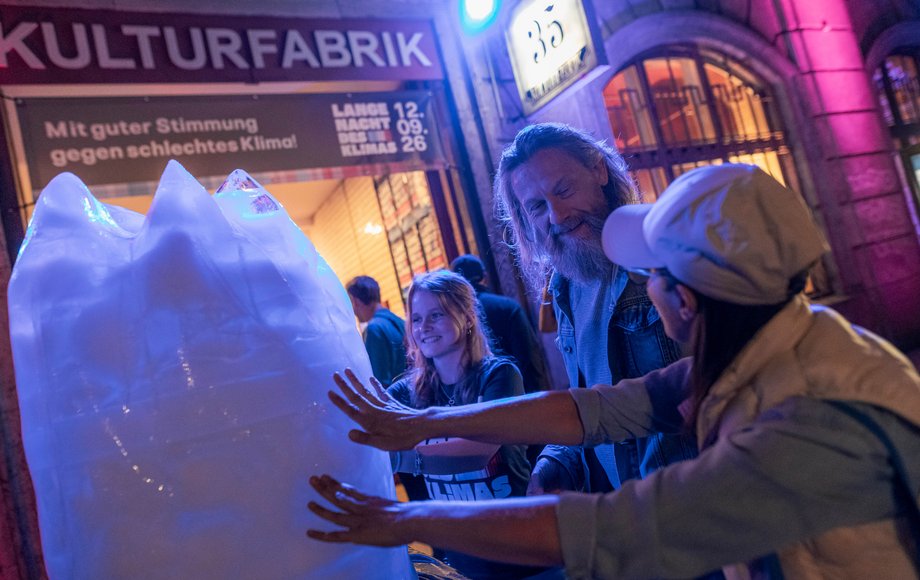 Um einen Eisblock stehen drei Menschen.