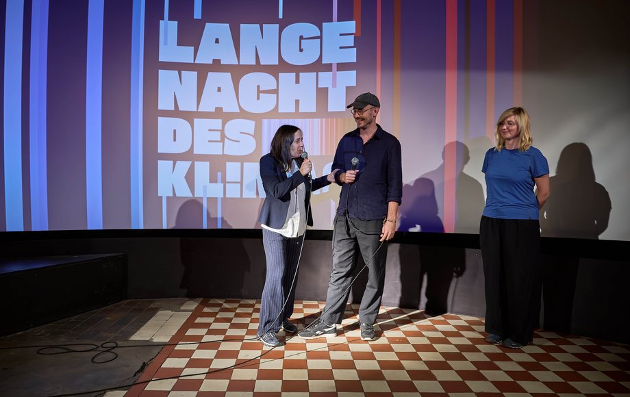 Talkrunde von zwei Frauen und einem Mann vor Warming-Stripes-Backdrop.