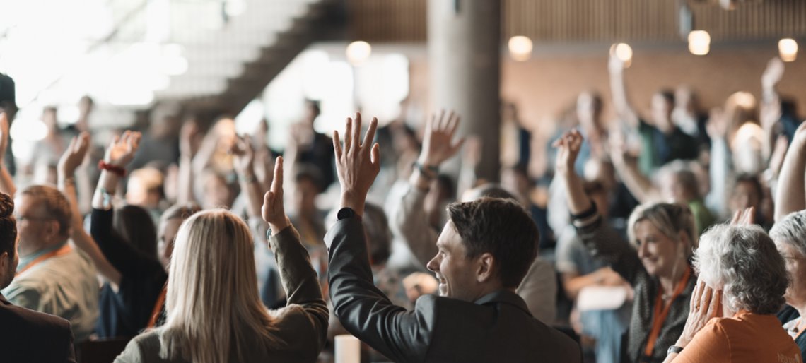 Eine große Versammlung, der Großteil der abgebildeten Menschen meldet sich