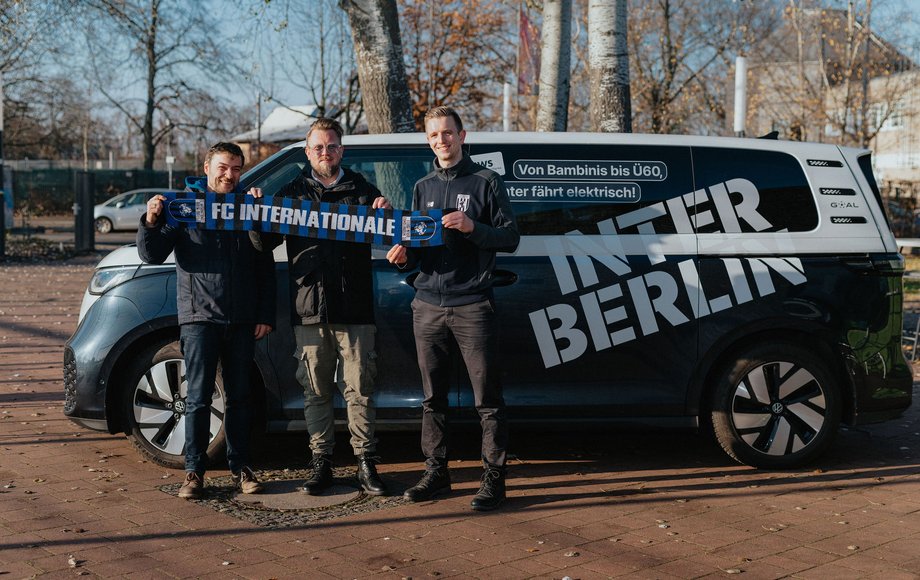 Vier Männer stehen nebeneinander vor einem VW-E-Bus und halten einen Fanschal des FC Internationale Berlin