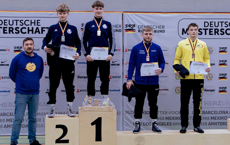 Siegertreppchen mit den Gewinnern, das von zwei weiteren Personen flankiert wird