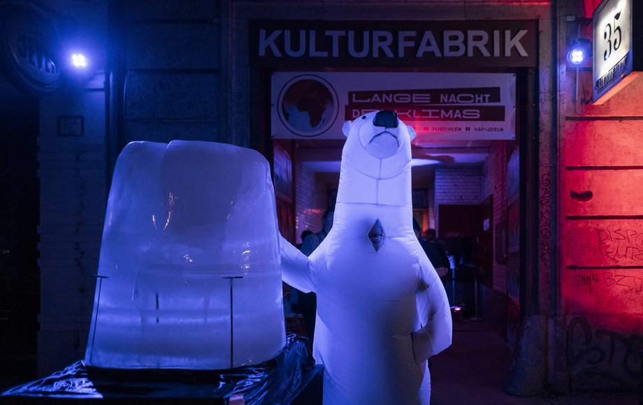 Vor einem bunt ausgeleuchteten Eingang eines alten Fabrikgebäudes steht ein Eisberg und eine Eisbärenfigur.