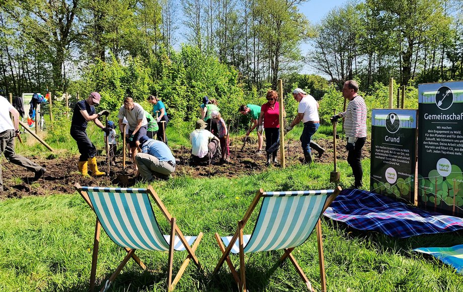 Szenerie auf einer Wiese. Im Vordergrund des Bildes stehen zwei Liegestühle, im Hintergrund gräbt eine Gruppe von Menschen den Boden auf, am rechten Bildrand ist ein Zaun mit Info-Transparenten