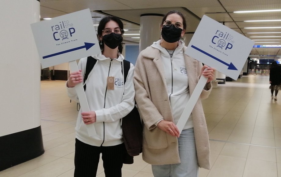 Zwei junge Mädchen halten Wegweiserschilder am Bahnhof mit Aufschrift „Rail to the COP“